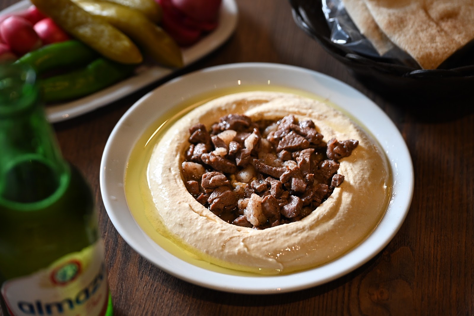 Hummus topped with spiced meat and pine nuts, pickles behind