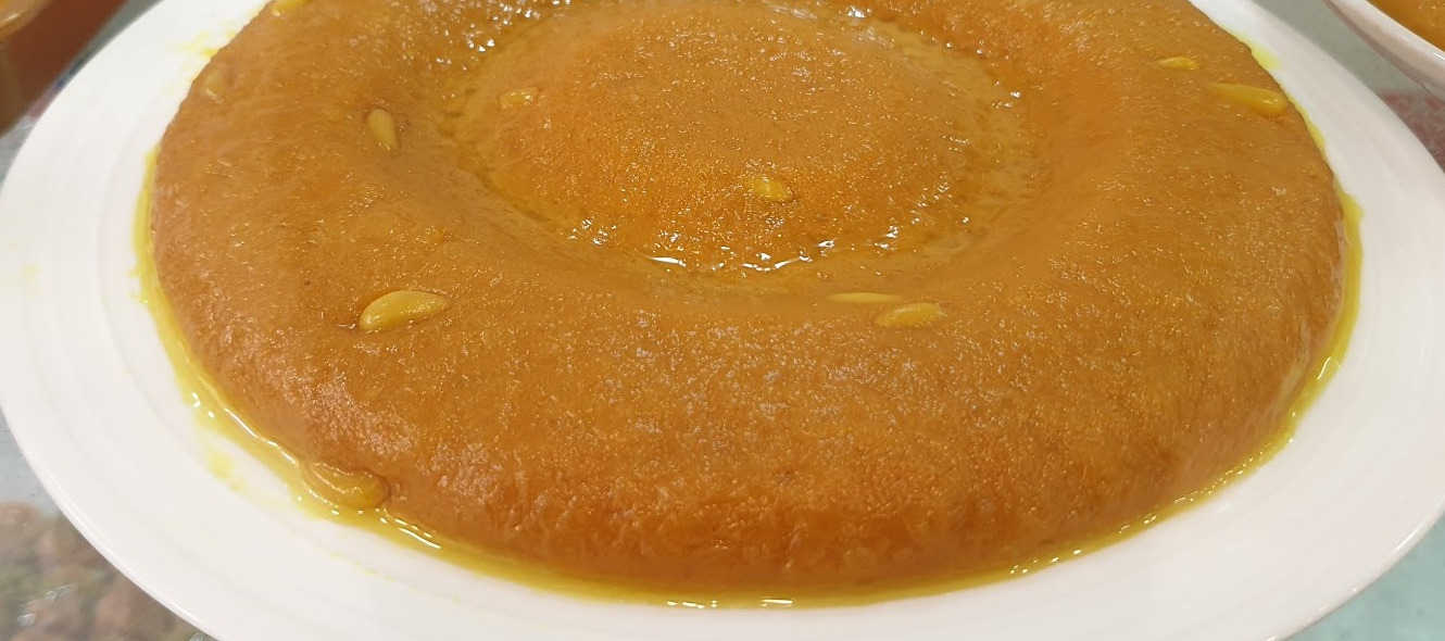 A whole mfatqa, turmeric semolina pudding with pine nuts, on a white plate at Al Makari Sweets