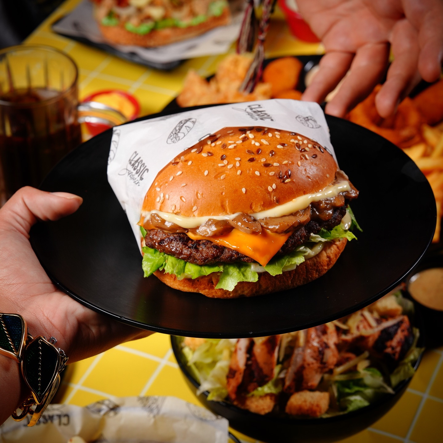 A burger on a black plate held out over the yellow tables