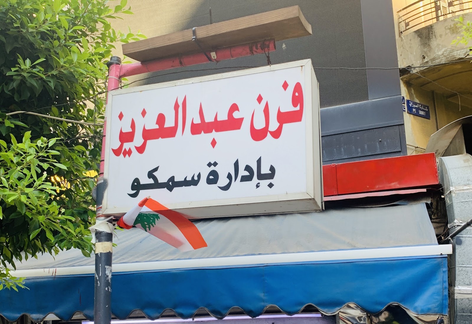The Furn Abdel Aziz lightbox sign above the blue awning, Lebanese flag beside it