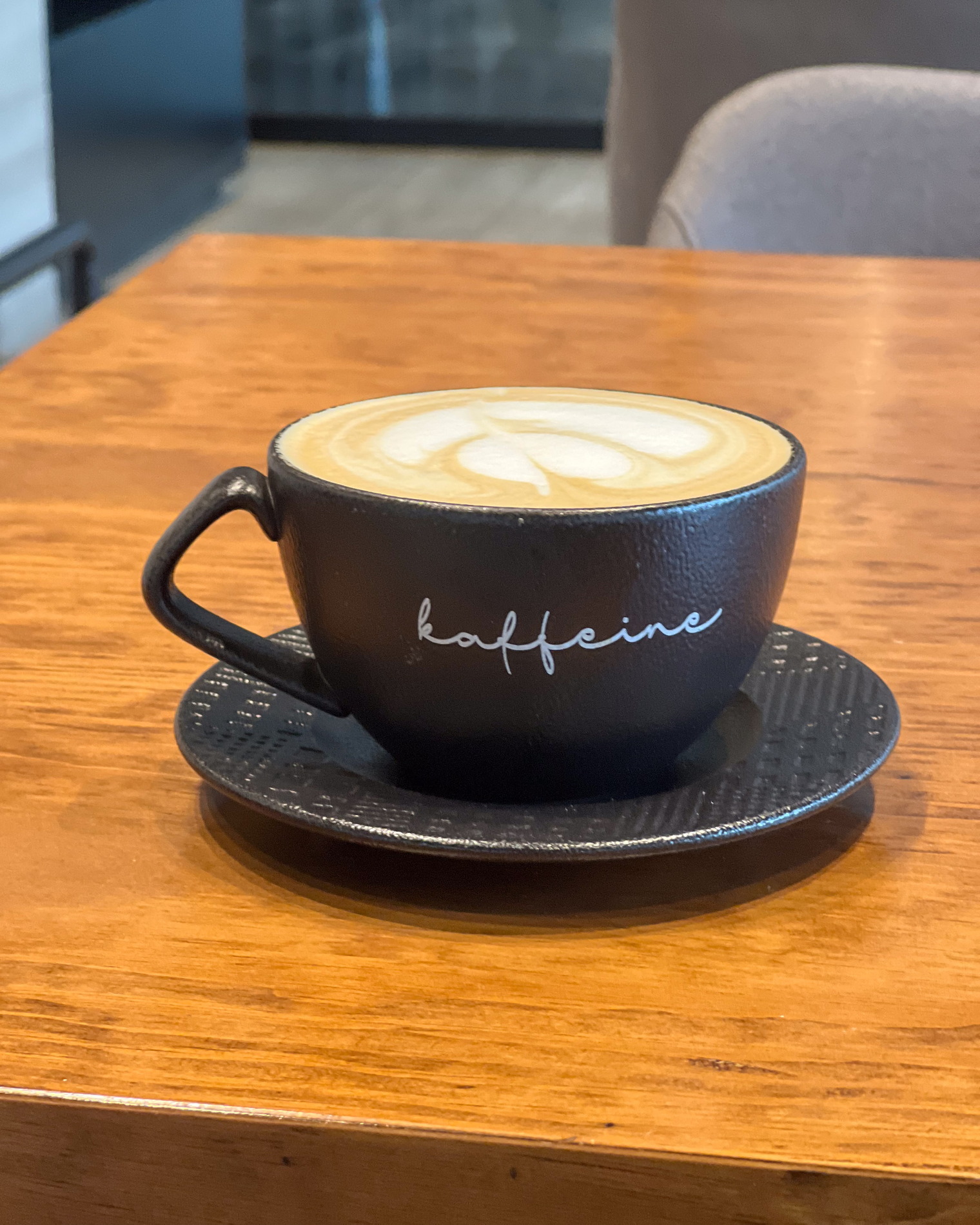 A cappuccino with poured latte art in a black cup marked kaffeine, on the wooden table