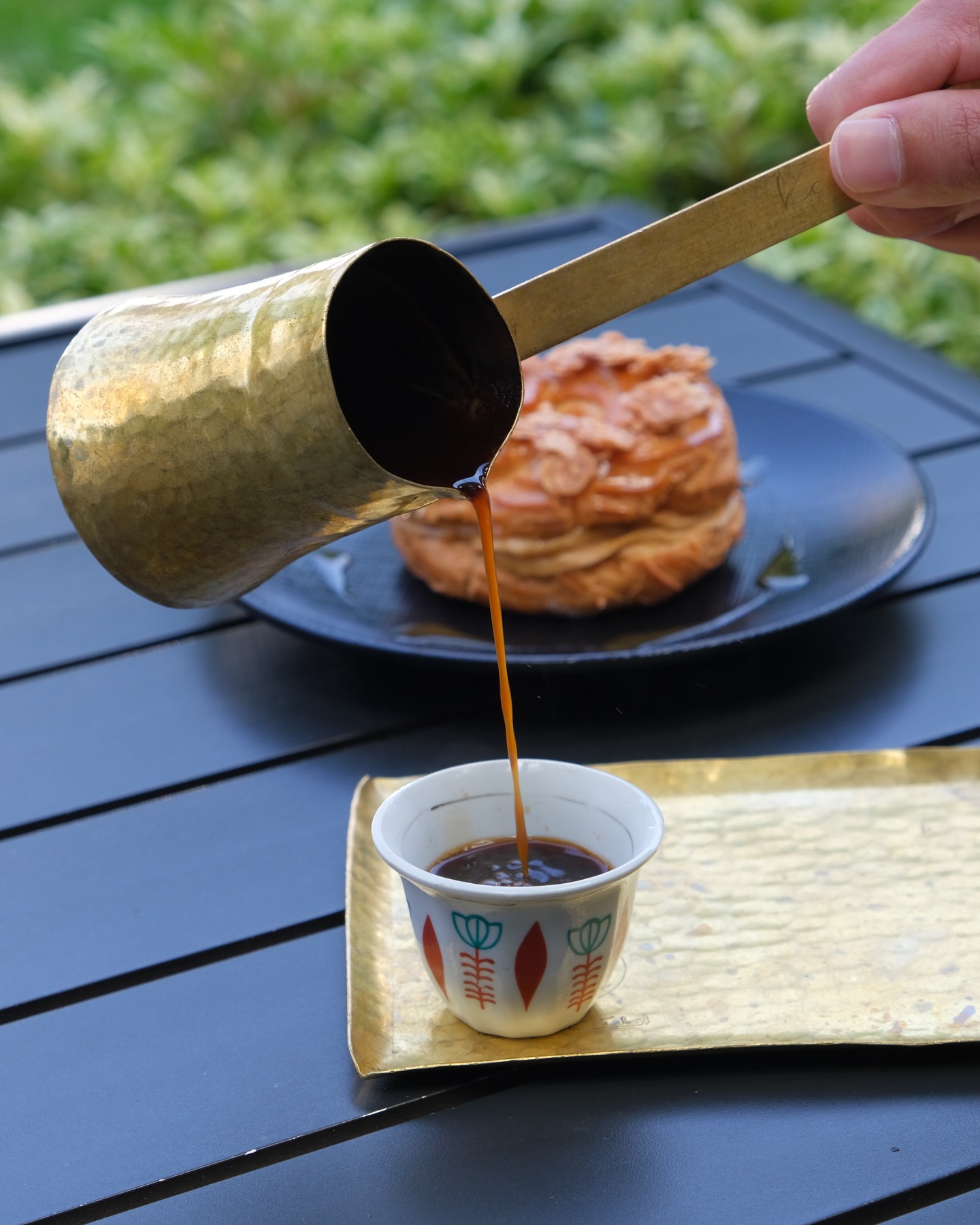Lebanese coffee poured from a brass rakwa into a small patterned cup beside a pastry