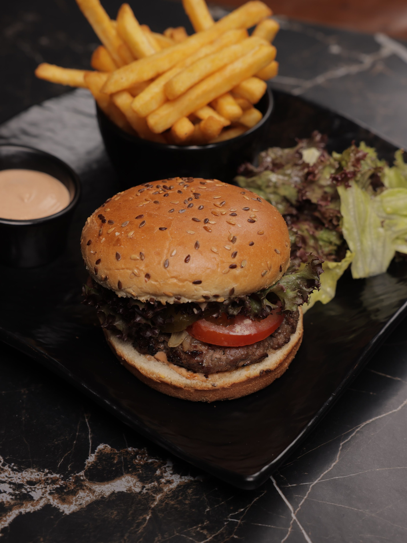 A sesame bun burger with fries and sauce on a black marble board