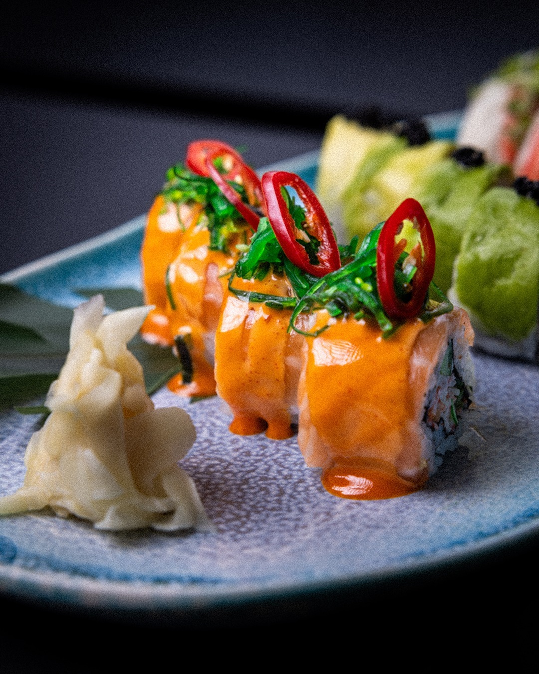 A spicy salmon roll topped with chilli and herbs on a speckled blue plate