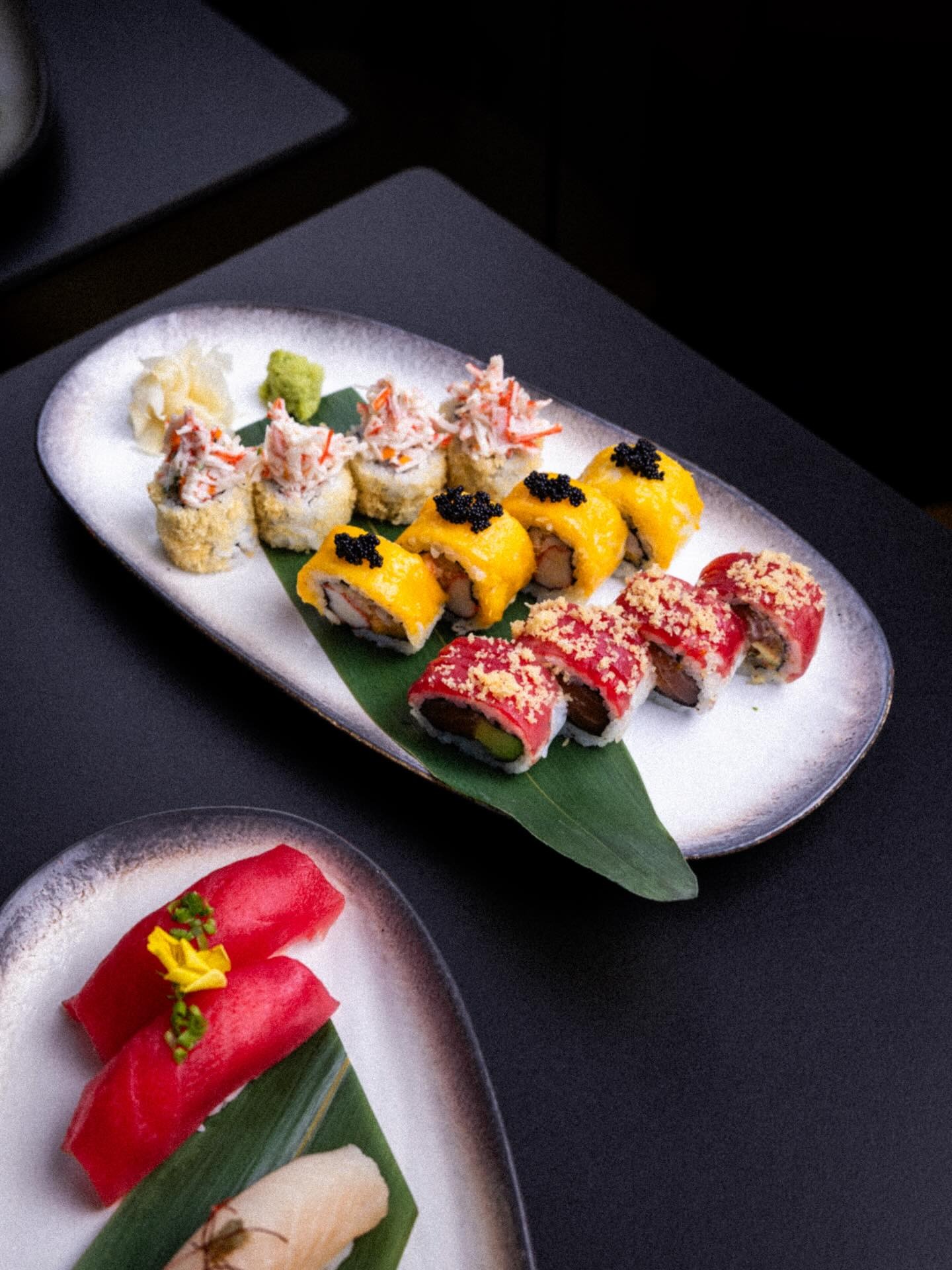 A long white plate of rolls beside nigiri cut thin on a green leaf