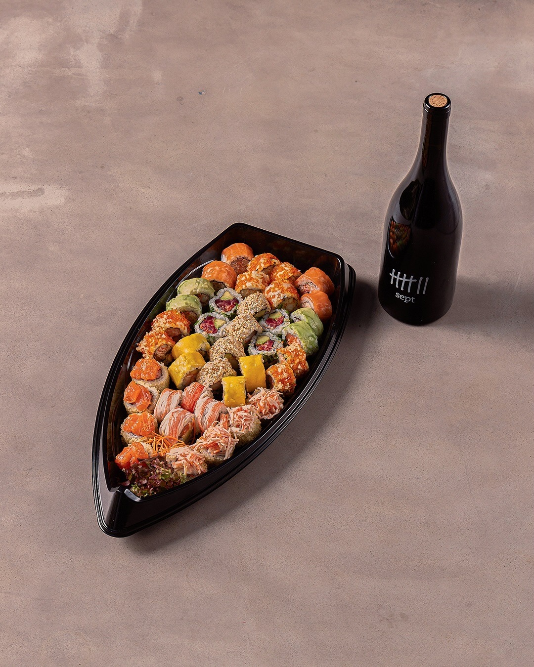A black boat platter packed with rows of rolls beside a dark bottle