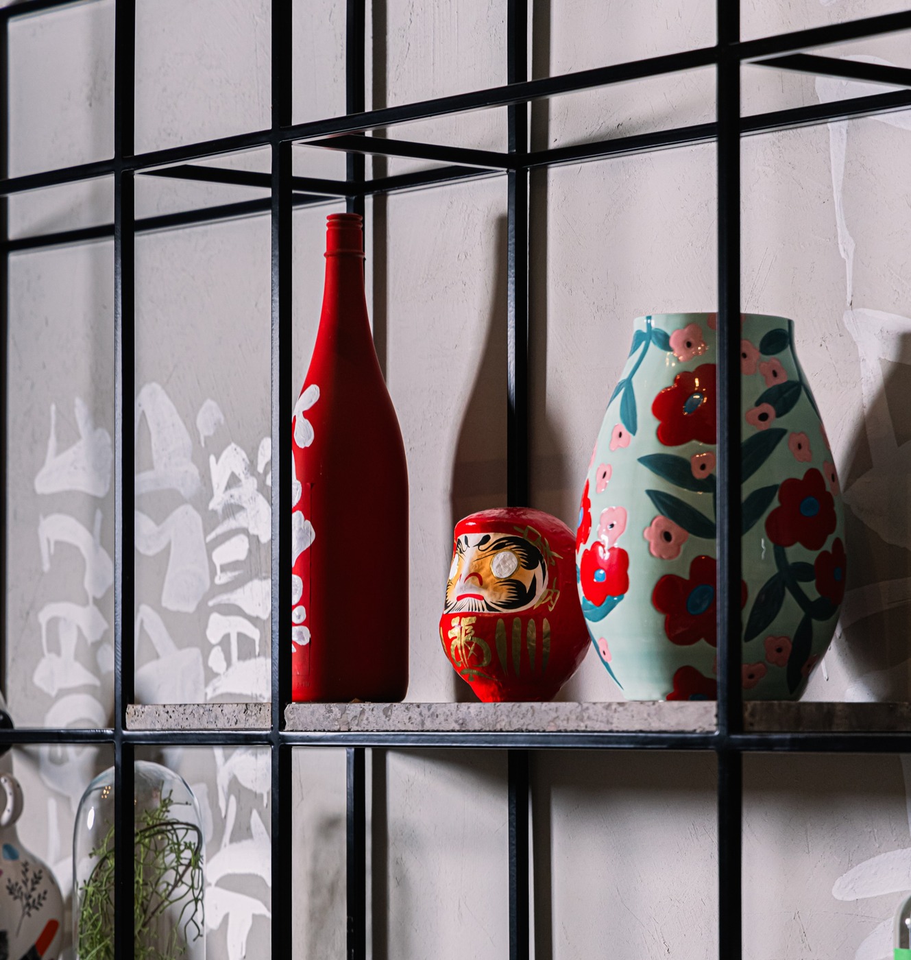 A red sake bottle, a daruma and a painted flower vase on a black grid shelf