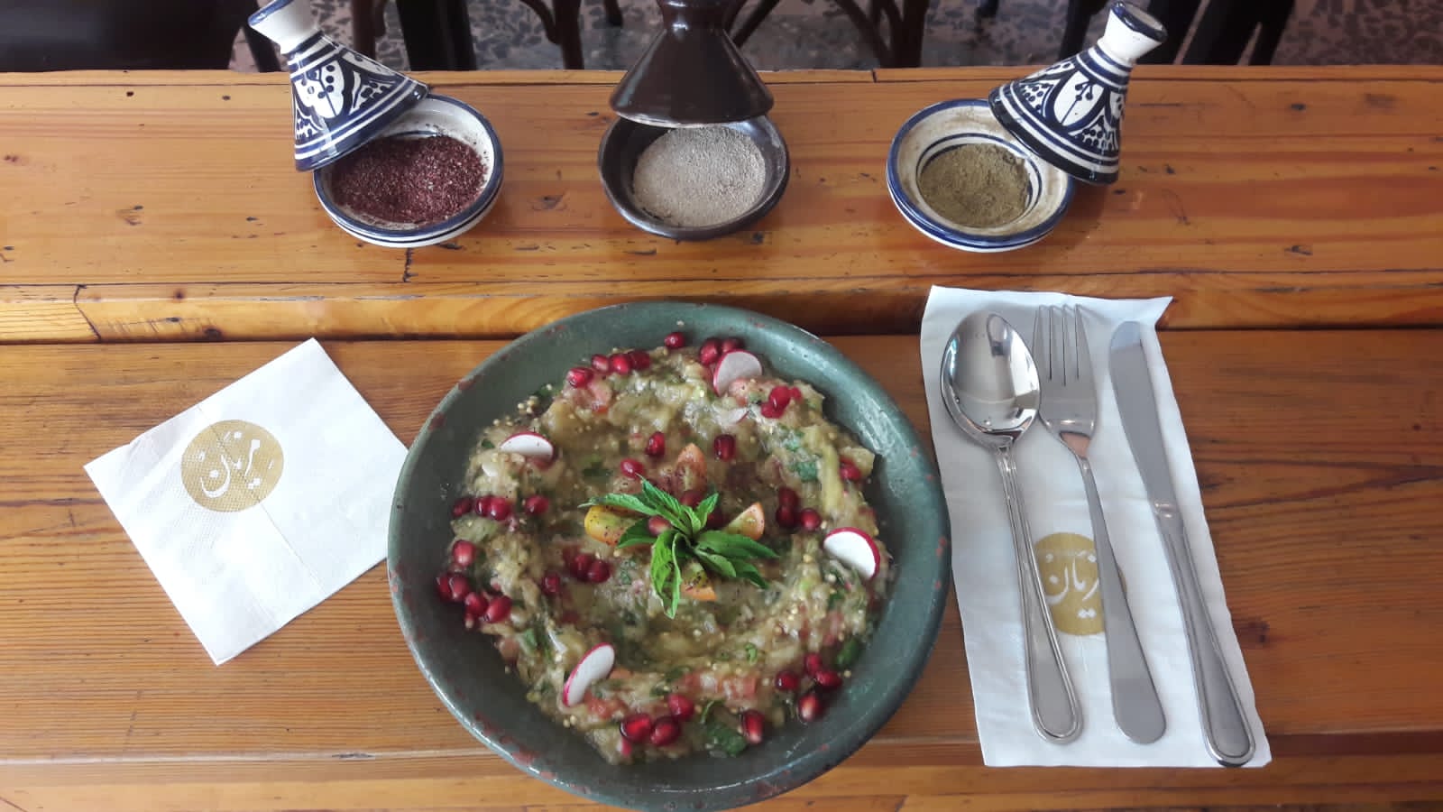 Mezyan's table from above: the moutabbal under pomegranate on the timber, the cutlery, and the napkins stamped in gold