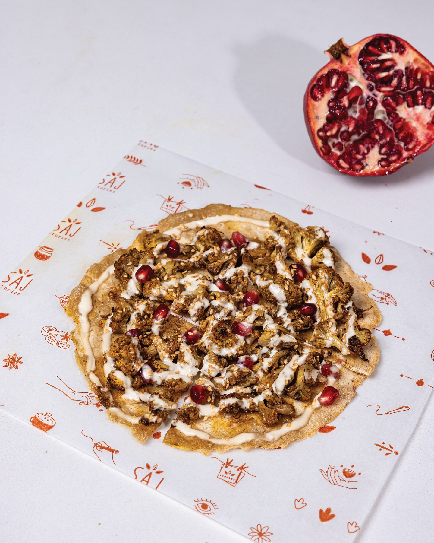 A saj under a tahini swirl and pomegranate seeds, a split pomegranate beside it