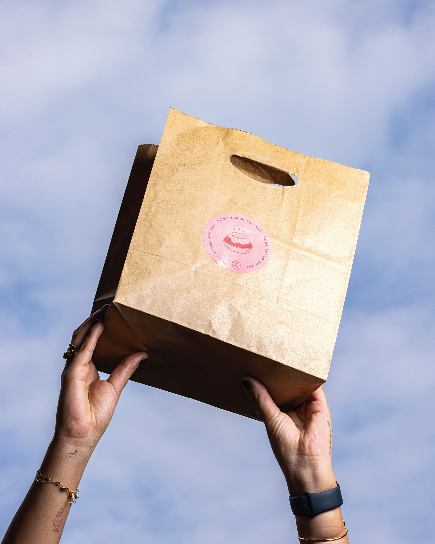 A stamped kraft Saj Stories bag held up against the sky